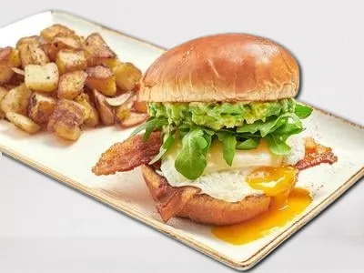 Elevated Egg Sandwich from First Watch, featuring crispy bacon, an over-easy cage-free egg, Gruyere cheese, fresh smashed avocado, mayo, and lemon-dressed arugula on a brioche bun.