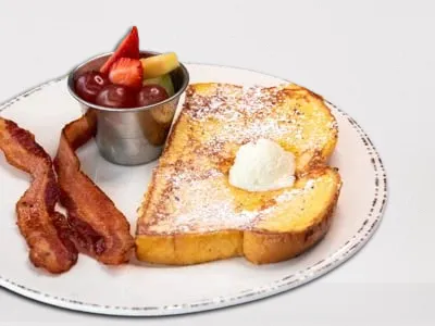 Kids French Toast from First Watch, featuring fluffy brioche bread dusted with powdered cinnamon sugar, served with fresh seasonal fruit and your choice of bacon or sausage.